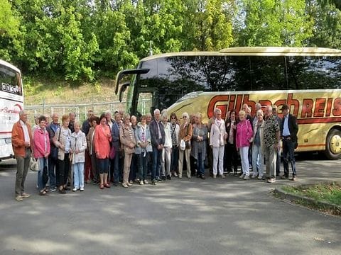 Eine Reisegruppe steht an einem sonnigen Tag vor einem gelben Reisebus, im Hintergrund Bäume.